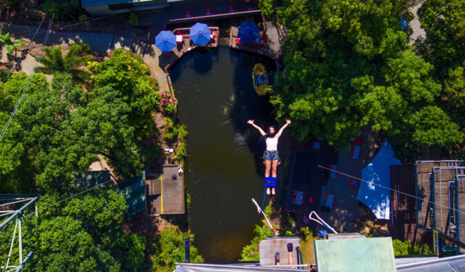 Woman bungee jumping over water surrounded by trees arms out facing up to camera