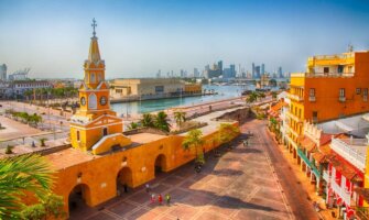 A view over an expansive plaza surrounded by bright orange historic buildings with the harbor and modern skyscrapers in the background in Cartagena, Colombia