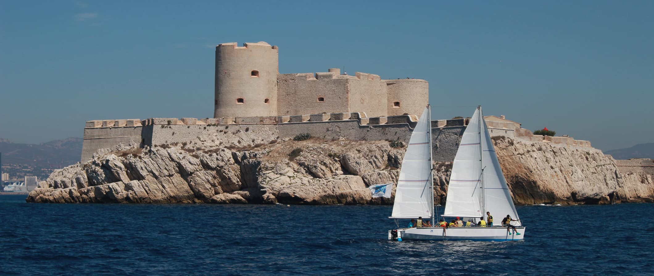 A small sailboat filled with passengers passing in front of Château d’If, a fortress off the coast of Marseille